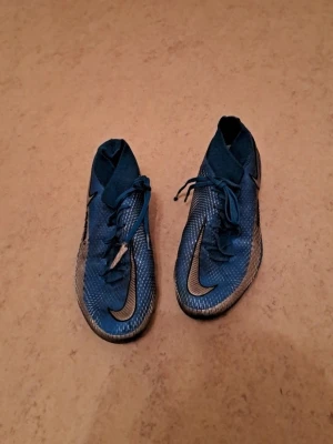 Nike Phantom Vision blå fotbollsskor - Säljer ett par blå Nike Phantom Vision fotbollsskor med silverfärgade detaljer och snörning. Skorna har högre strumpkrage för extra stöd runt ankeln och mönstrad ovansida för bättre bollkontroll. Sulan har fasta dobbar som passar perfekt för spel på gräs. I mycket bra skick. Säljs för 150 kr. Tveka inte att kontakta om du är intresserad eller har några frågor! Först till kvarn dom gäller!