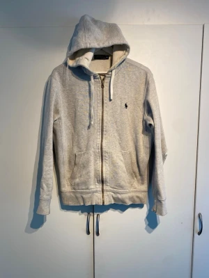 Ralph Lauren Zip Hoodie  - Säljer en Ralph Lauren Zip Hoodie i ljusgrå färg. Storlek M. Använt skick med flera defekter, därav priset. Hål vid armbågar. Flera lösa trådar med mera. Nypris 1800kr