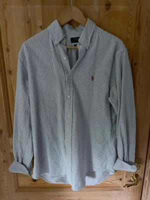 Randig skjorta från Polo Ralph Lauren - Klassisk långärmad skjorta från Polo Ralph Lauren i vitt och blått med tunna ränder. Skjortan har knappar framtill, klassisk krage och den ikoniska broderade loggan på bröstet. Tillverkad i mjuk bomull och har en avslappnad passform.
