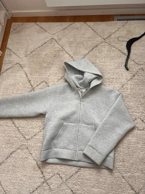 Grå zip hoodie i scuba-material - Säljer en stilren grå hoodie i scuba-material med dragkedja framtill och stor huva. Hoodien har två rymliga fickor på magen och breda muddar vid ärmslut och nederkant. Perfekt för en avslappnad och trendig look. Passar folk med S och M nypris 650 