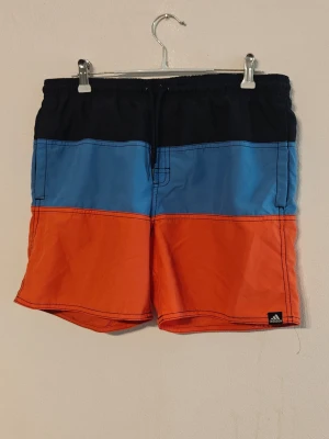 Adidas badshorts  - Säljer ett par snygga badbyxor från Adidas i färgblock med svart, blått och orange. De har resårmidja med dragsko och kortare ben. Perfekta för stranden eller poolen. Adidas-logga på benet och tillverkade i lätt, snabbtorkande material. Storlek XL för barn.