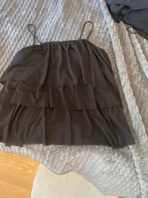 Svart volangtop med tunna band - Säljer en svart topp med tunna axelband och flera lager volanger. Toppen har en luftig och flowy passform, perfekt för dig som gillar en lite mer lekfull stil. Materialet känns lätt och svalt, och toppen är ärmlös med en rak skärning upptill.