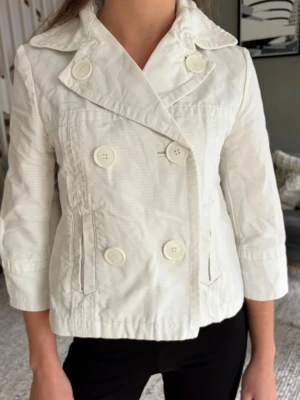 Vit kort trenchcoat med stora knappar - Snygg vit trenchcoat med dubbelknäppning och stora matchande knappar. Jackan har trekvartsärmar, bred krage och två fickor framtill. Tillverkad i ett lätt tyg som passar perfekt till vår och sommar. Stilsäker och enkel att matcha till olika outfits.