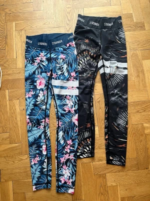 Två par mönstrade leggings från Stronger - Säljer två par tights från Stronger. Ena paret har ett tropiskt blommönster i blått, rosa och vitt, medan det andra har ett svart mönster med inslag av vitt och orange. 