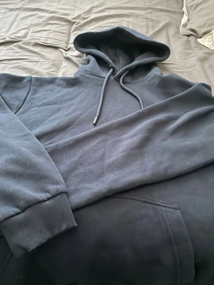Mörklblå Jack&Jones hoodie - En enkel och stilren mörkblå hoodie med huva och dragsnören. Hoodien har en klassisk känguruficka framtill och ribbade muddar vid ärmslut och nederkant. Perfekt för en avslappnad och bekväm look. Aldrig använd