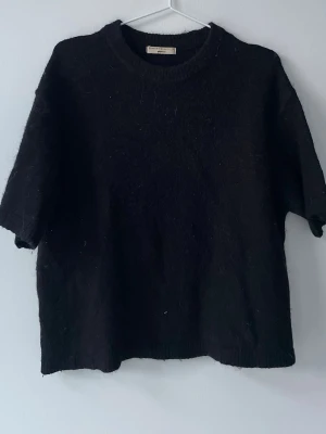 Svart stickad tröja från Gina Tricot - Svart stickad tröja från Gina Tricot med korta ärmar och rund halsringning. Tröjan har en enkel och stilren design, tillverkad i ett mjukt och fluffigt material som känns skönt mot huden. Perfekt för lager-på-lager och en avslappnad look.