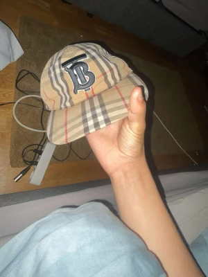 Burberry TB keps - Säljer en beige keps med klassiskt rutigt mönster och stor svartvit B-logga framtill. Kepsen har justerbar rem bak med tryckknappar och är tillverkad i ett lätt tygmaterial. Perfekt för dig som vill sticka ut med en trendig accessoar.