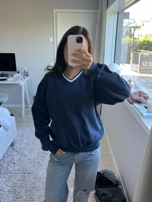 Mörkblå sweatshirt med V-ringning - Mörkblå stickad tröja med V-ringad hals och vita detaljer runt kragen. Tröjan har en avslappnad passform, långa ärmar och ribbade muddar vid ärmslut och nederkant. Perfekt för en chill och stilren look.