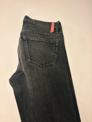 Acne Jeans - Säljer ett par svarta jeans med rak passform och klassisk femficksdesign. Jeansen har en diskret tvättad vintage-look och en svart patch med röd detalj i linningen. Perfekta för en avslappnad och trendig stil.