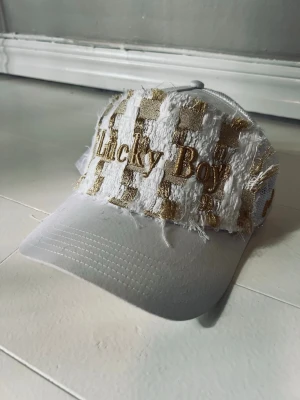 Vit keps j dauphin  - Säljer en unik vit keps med slitna detaljer och guldiga broderier. Framtill står det 'Lucky Boy' och på sidan finns ytterligare broderad text. Kepsen har mesh på baksidan för extra ventilation och en cool streetwear-look.