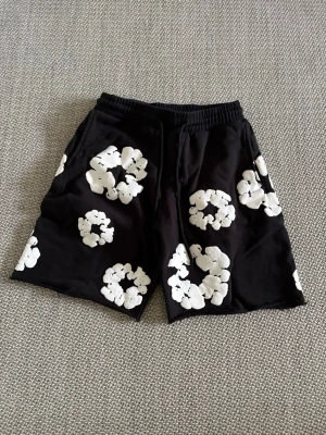 Svarta shorts med vita blommor Denim Tears - Säljer ett par svarta shorts från Denim Tears med stora vita blommönster över hela plagget. Shortsen har elastisk midja och sidofickor, perfekta för en chill och trendig look. Materialet känns mjukt och bekvämt, perfekt för varma dagar.