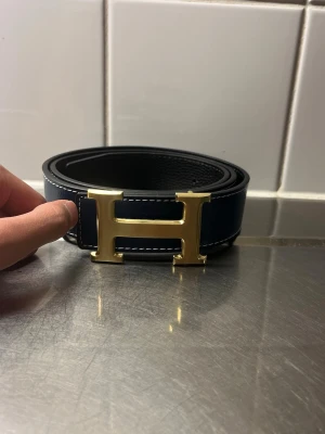 Svart bälte med guldspänne - Snyggt svart bälte med ett stort H-format spänne i guld. Bältet har vita sömmar längs kanterna och är gjort i skinn. Perfekt accessoar för att lyfta din outfit.