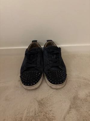 Marin blåa sneakers med nitar - Säljer ett par marin blåa sneakers med coola marin blåa nitar på tån och klassisk röd sula. Skorna har snörning och en låg profil som passar till många olika stilar. Materialet ser ut att vara mocka eller liknande tyg. Stolek 41 passar 42 
