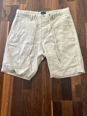 Beige linneshorts från Gant med snörning - Snygga beige shorts från Gant i linne med snörning i midjan och knappstängning. De har en avslappnad passform och praktiska sidofickor. Perfekta för varma dagar och ger en clean och stilren look.
