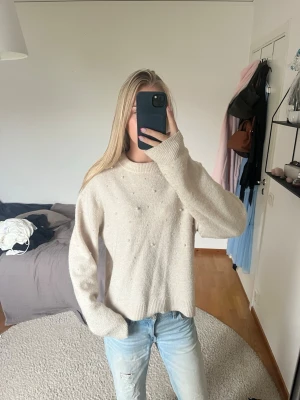 Beige stickad tröja med pärldetaljer - Säljer en beige stickad tröja med små pärldetaljer framtill. Tröjan har rund halsringning, långa ärmar och en avslappnad passform. Perfekt att matcha med jeans för en enkel och snygg look.