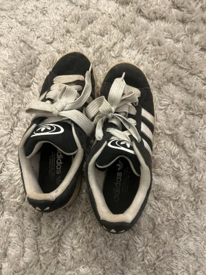 Svarta Adidas sneakers med vita ränder - Klassiska svarta Adidas sneakers med vita ränder och vit sula. Skorna har Adidas logga på plösen och hälen, samt vita skosnören. Ovandelen är i mockaliknande material och modellen är lågskuren för en avslappnad look.