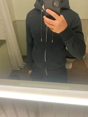 Svart hoodie med dragkedja från GANT - Snygg svart hoodie från GANT med dragkedja framtill och broderad logga på bröstet. Huva med dragsko och ribbade muddar vid ärmslut och midja. Perfekt för en avslappnad och stilren look.
