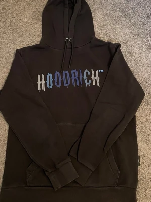 Hoodrich hoodie - Svart hoodie med stor huva och dragsko. Framtill finns ett färgglatt HOODRICH-tryck i blått, vitt och beige. Hoodien har en klassisk känguruficka och ribbade muddar vid ärmslut och nederkant. Perfekt för en chill och streetinspirerad look.