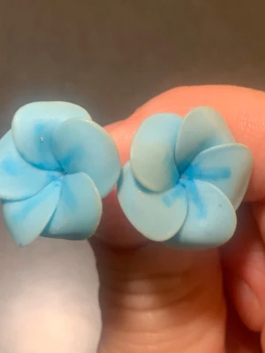 Blå blomformade örhängen - Säljer ett par unika örhängen i form av blå blommor. De är gjorda i ett lätt material, troligen plast eller lera, och har en mjuk, pastellblå färg med lite mörkare blå detaljer. Perfekta för dig som vill ha något färgglatt och annorlunda i öronen. Ca 2cm långa och 1,5cm breda, använda vid enstaka tillfällen och är i väldigt bra skick 
