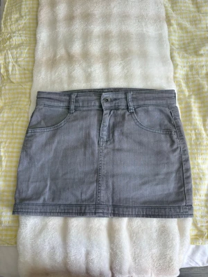 Grå kort jeanskjol med fickor - Snygg grå kort jeanskjol med klassisk femficksdesign, bälteshällor och knappstängning fram. Kjolen har två fickor fram och två bakfickor med dekorativa sömmar. Perfekt till vardags för en avslappnad och trendig look.