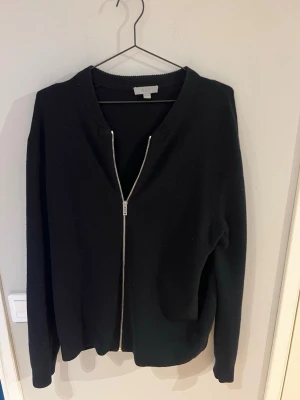 Svart zip-kofta från COS - Stilren svart kofta från COS med dragkedja framtill. Koftan har en enkel design med ribbad krage och långa ärmar. Perfekt att slänga på sig över en t-shirt eller skjorta för en clean look.