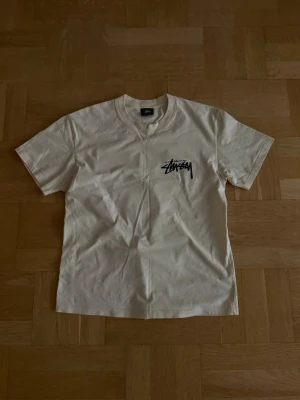 Vit Stüssy t-shirt med logga - Vit t-shirt från Stüssy med klassisk svart logga tryckt på bröstet. T-shirten har rund halsringning och korta ärmar. Perfekt för en avslappnad streetwear-look.