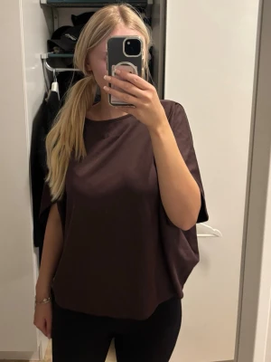 Brun lös satinblus med kort ärm - Säljer en mörkbrun blus i satin med lös passform och korta ärmar. Toppen har en enkel, rund halsringning och ett snyggt, minimalistiskt uttryck. Perfekt för dig som gillar stilrena och bekväma plagg. Storleken är XS/S