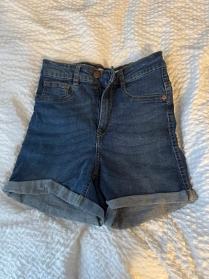 Blå högmidjade jeansshorts Molly - Säljer ett par blå högmidjade jeansshorts från Gina Tricot, modell Molly. Klassisk femficksdesign, uppvikta benslut och knappgylf. Perfekta till sommaren och lätta att matcha med olika toppar.