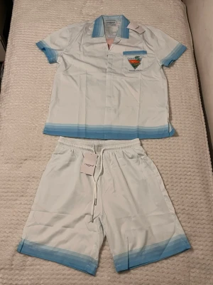 Casablanca Tennis Club set i vitt och blått - Säljer ett stilrent set från Casablanca med kortärmad skjorta och shorts i vitt med ljusblå detaljer. Skjortan har tryck med 'Casablanca Tennis Club' och färgglad grafik både fram och bak. Materialet känns lätt och svalt, perfekt för sommaren.