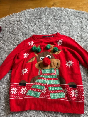 Röd jultröja med renmotiv och tofsar - Rolig röd stickad jultröja med ett stort renmotiv framtill, dekorerad med gröna och röda tofsar som ger en 3D-effekt. Tröjan har vita snöflingor och mönster längs nederkanten samt långa ärmar och rund hals.