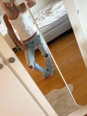 Ljusblå slitna bootcut jeans - Säljer ett par ljusblå jeans med bootcut-modell och flera slitningar och hål på benen för en trendig, avslappnad look. Jeansen har klassisk femficksdesign och låg midja. Perfekta för dig som gillar en edgy och casual stil. Är från Gina Tricot men gjort hålen och det själv 