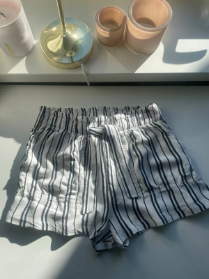 Randiga shorts med knyt från H&M - Snygga vita shorts från H&M med svarta ränder och knyt i midjan. De har elastisk midja och är gjorda i mjuk bomull, perfekta för varma dagar. Stilen är avslappnad och passar till allt.