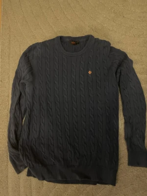 Mörkblå Morris kabelstickad tröja - OBS står XL men passar L/M mest L. Mörkblå kabelstickad tröja med rund halsringning och ribbade muddar. Tröjan har ett litet broderat emblem på bröstet och är långärmad. Perfekt för lager-på-lager och passar till många olika stilar. OBS den har ett litet hål som har sytt för inte för tydligt 