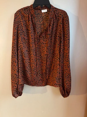 Rostfärgad, leopardmönstrad blus från VILA - Snygg rostfärgad blus från VILA med svart leopardmönster. Blusen har långa ärmar med resår vid ärmslut och en lös passform. Fin knytning i halsringningen. Mycket användbar!