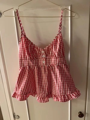 brandy melville topp - Supersöt linnetopp med smala axelband i rödvitt rutigt mönster. Toppen har knappar framtill, volangkant nedtill och smockad rygg för extra komfort. Perfekt för varma dagar och ger en riktigt somrig vibe.