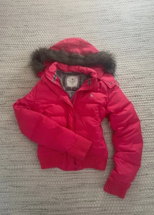 Rosa pufferjacka från Abercrombie & Fitch - Snygg rosa pufferjacka från Abercrombie & Fitch med avtagbar huva och fuskpälsdetalj. Jackan har ribbade muddar, dragkedja och knappar framtill samt två fickor. Perfekt för kalla vinterdagar och riktigt trendig look.