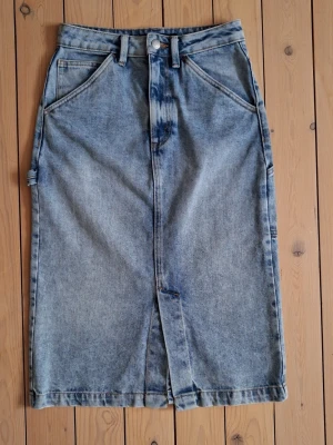 Lång jeanskjol med fickor - Lång ljusblå jeanskjol från Bik Bok. Kjolen har en rak passform och slits fram och baktill för extra rörelsefrihet. Kjolen är i dint skick men har en luten nötning på baksidan (se bild 7).              Midja ca 66 cm (ca 33 cm raktöver).            Längd ca 66 cm.