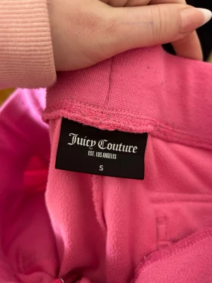 Rosa byxor från Juicy Couture - Säljer en ikonisk rosa byxor från Juicy Couture i mjukt material. Byxorna har broderad JC-logga på bröstet och klassisk bakfickor. Perfekt för dig som vill ha en trendig och bekväm look.