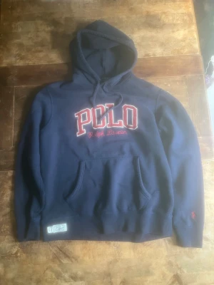 Marinblå hoodie från Polo Ralph Lauren - Snygg marinblå hoodie från Polo Ralph Lauren med stor röd och vit POLO-logga broderad på bröstet. Klassisk känguruficka framtill och dragsko i huvan. Mjuk bomullskänsla och diskret logga på ärmen. Perfekt för en chill och sportig look. I princip nyskick då den bara använts kort inomhus i hemmet ett par gånger då den provats. Har sedan blivit liggandes i garderoben. 