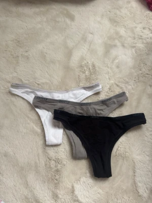 3-pack SKIMS stringtrosor i bomull - Säljer ett 3-pack stringtrosor från SKIMS i färgerna svart, grå och vit. Trosorna är tillverkade i mjuk bomull med elastisk midja där SKIMS-loggan syns. Perfekta för dig som gillar bekväma och stilrena underkläder.