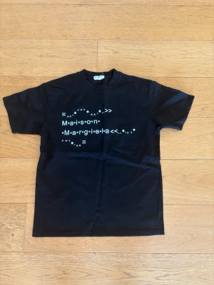 Maison Margiela svart t-shirt - Svart t-shirt från Maison Margiela med vit grafisk text på bröstet. Klassisk rund halsringning och korta ärmar. Tillverkad i mjuk bomull för en bekväm känsla och stilren look.