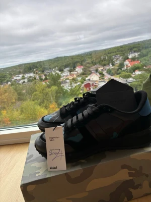 Valentino Rockrunners - Snygga svarta sneakers från Valentino med camo-mönster i blått och svart. Skorna har svarta snören, robust sula och detaljer i läder och textil. Perfekta för dig som vill sticka ut med en exklusiv och trendig look. Varan är köpt från miinto för 3499kr Man får även med tillbehör såsom box, extra snören, äktighetscertifikat och en valentino påse. Jag säljer dessa skor pga att dem var för stora när jag köpte dem. Dem är helt oandvända. Hör av dig om du har några frågor 😀