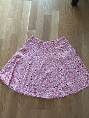 Rosa blommig kortkjol XS - Söt rosa kortkjol med vitt och svart blommönster. Kjolen har resår i midjan för extra komfort och en lätt utställd form. Perfekt för dig som gillar en feminin och avslappnad stil.