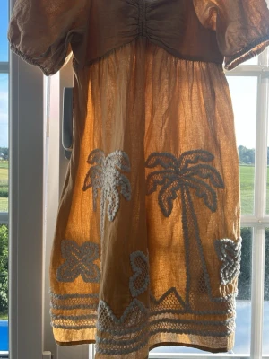 Beige kortklänning med broderi - Söt beige kortklänning i luftig bomull med puffärmar och broderade vita palmer och blommor nertill. Klänningen är i storlek 152 från Zara