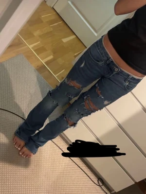 Blå slitna bootcut jeans - Säljer ett par blå bootcut jeans med kraftiga slitningar och hål på framsidan. Jeansen har låg midja och klassisk femficksdesign. Perfekta för en edgy och avslappnad stil.