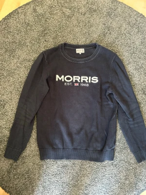 Mörkblå stickad tröja Morris - Mörkblå stickad tröja från Morris med rund halsringning och vit broderad logga samt texten 'EST. 1968' framtill. Tröjan har ribbstickade muddar vid ärmslut och nederkant. Perfekt för dig som gillar klassisk och stilren design.