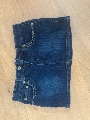 Mörkblå jeanskjol med kontrastsömmar - Snygg mörkblå jeanskjol i kort modell med kontrastsömmar och dekorativa fickor både fram och bak. Kjolen har knapp- och dragkedjestängning samt coola detaljer på fickorna. Perfekt för sommaren