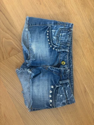 Blå jeansshorts med nitar - Snygga blå jeansshorts med slitna kanter och nitar på fram- och bakfickor. Shortsen är lågmidjade, korta, har klassiska fickor och bälteshällor. Perfekta för sommaren 