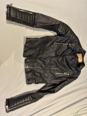 Svart skinnjacka med dragkedjor - Snygg svart skinnjacka i bikerstil med flera silverfärgade dragkedjor och detaljerade sömmar på ärmarna. Jackan har ståkrage med tryckknapp och två bröstfickor med dragkedja. Perfekt för en edgy look.
