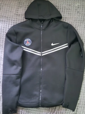 Svart PSG hoodie från Nike - Snygg svart hoodie med dragkedja från Nike och PSG-logga på bröstet. Två vita ränder går diagonalt över bröstet. Huva och långa ärmar, perfekt för dig som gillar sportig stil och vill representera Paris Saint-Germain.